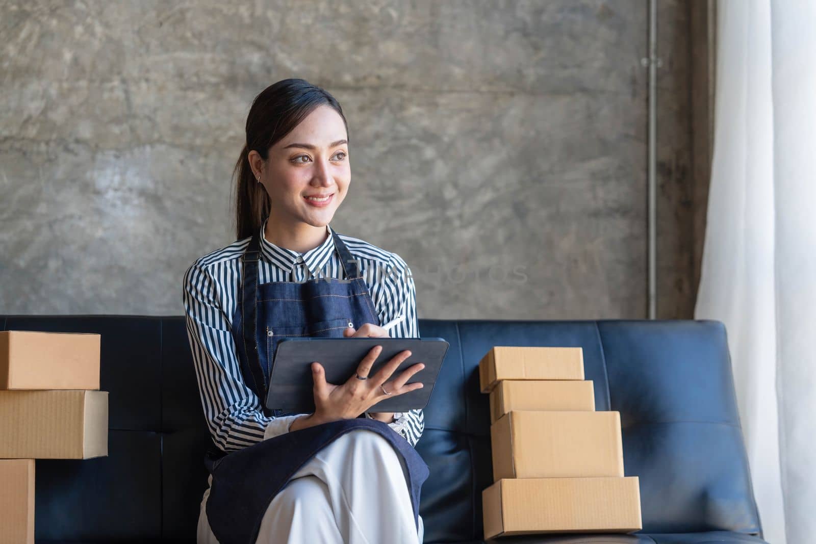 Young Asian business woman on sofa using laptop computer checking customer order online shipping boxes at home...