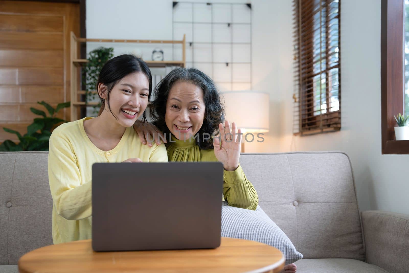 Happy adult granddaughter and senior grandmother having fun enjoying talk sit on sofa in modern living room, smiling old mother hugging young grown daughter bonding chatting relaxing at home together.