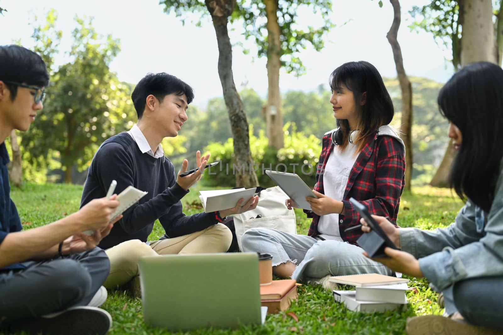 Happy university students talking, relaxing on campus lawn and working on laptop together. Education and lifestyle concept.