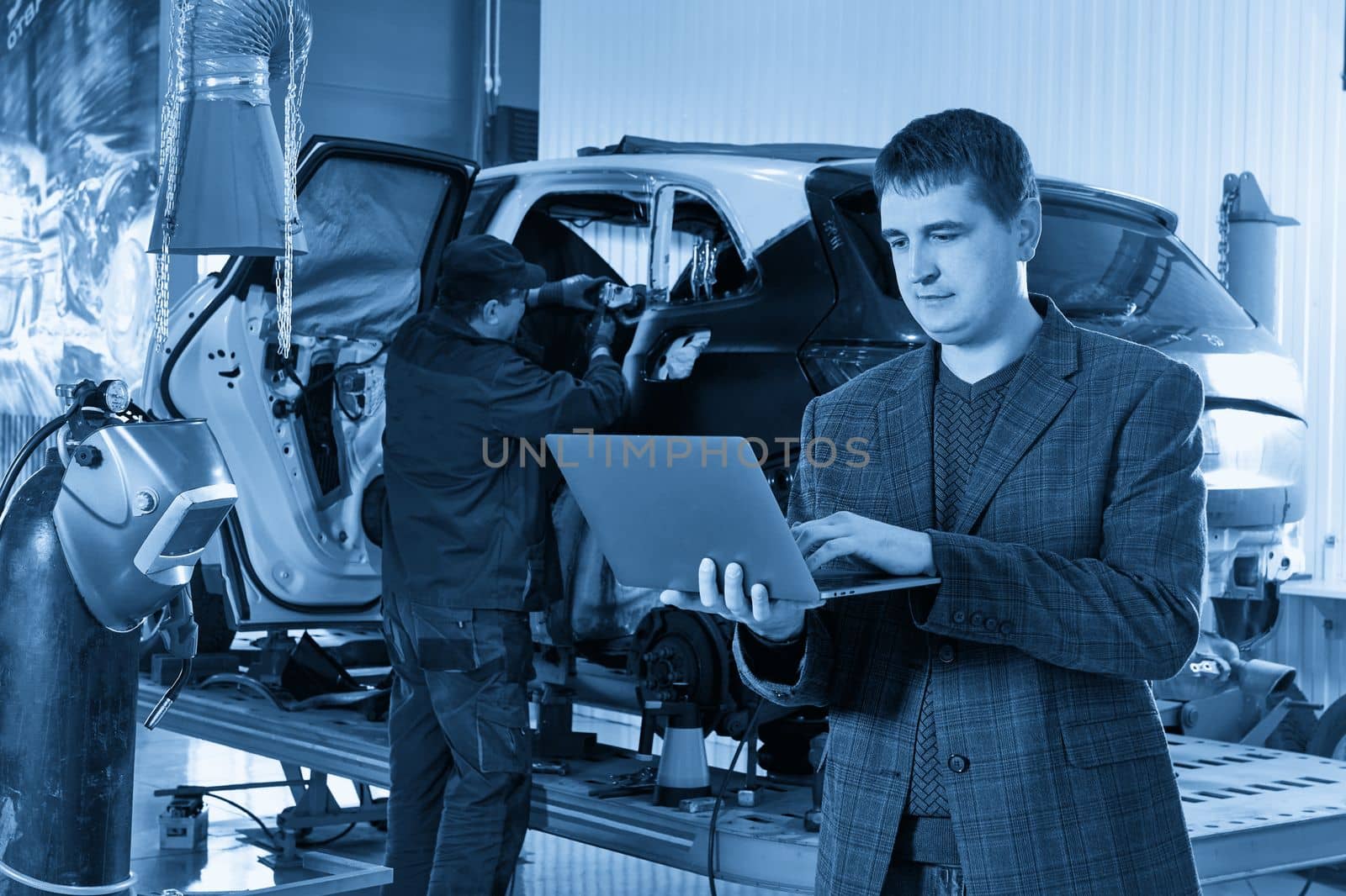 Manager holding a laptop while standing near car in auto repair shop with mechanic repairing car on background