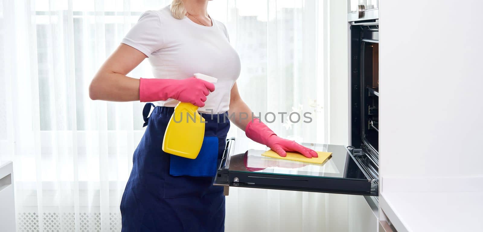 Young woman wearing gloves cleaning oven in the kitchen. Cleaning service concept by Mariakray