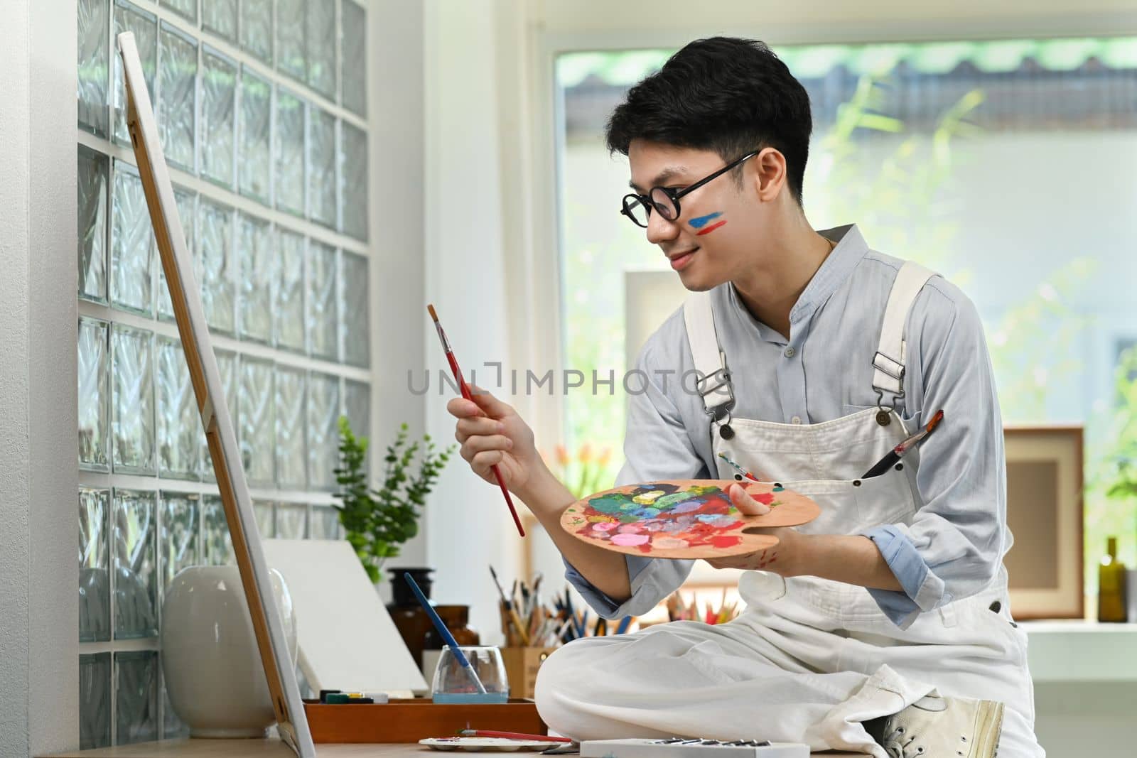 Smiling man artist sitting in front of canvas and painting picture with watercolor. Art and leisure activity concept.