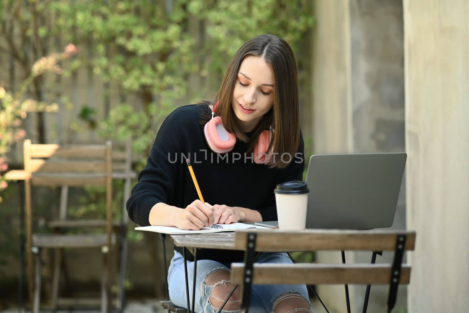 Pretty caucasian young woman writing down in notebook and using laptop while sitting at outdoor by prathanchorruangsak