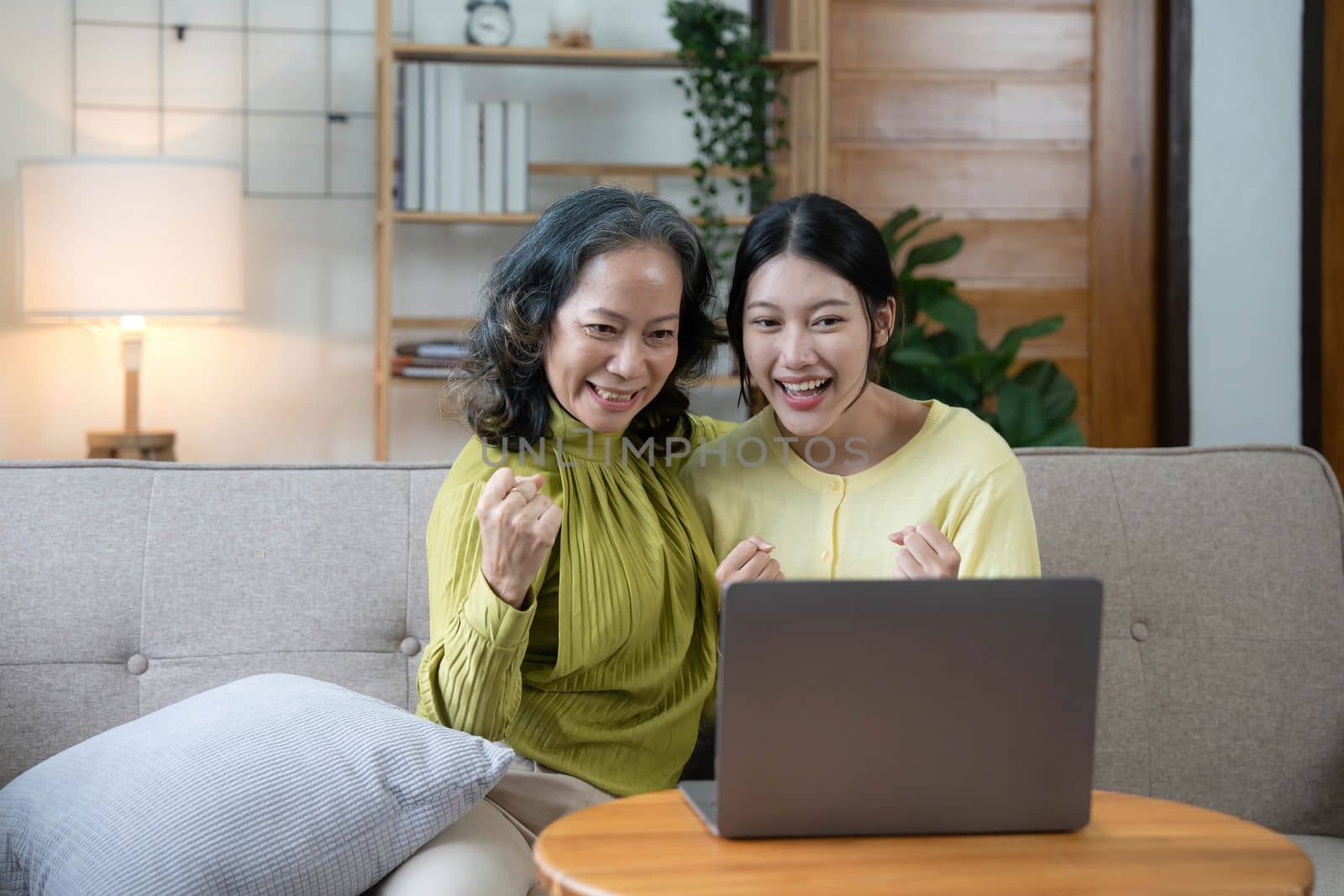 Happy adult granddaughter and senior grandmother having fun enjoying talk sit on sofa in modern living room, smiling old mother hugging young grown daughter bonding chatting relaxing at home together.