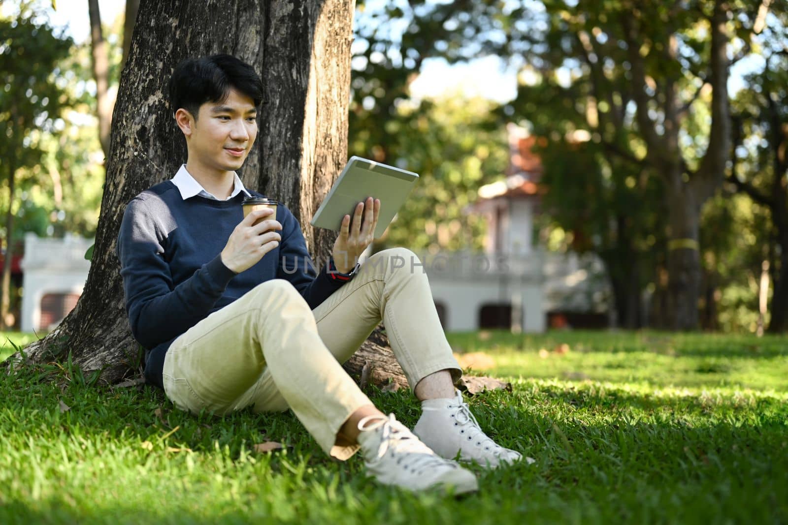 Pleased asian man drinking coffee and using digital tablet under tree on green grass at sunny beautiful day.
