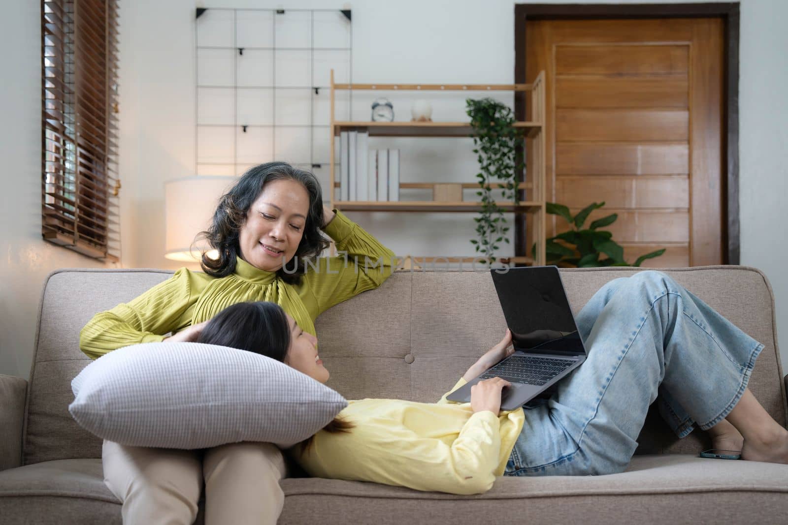 Happy adult granddaughter and senior grandmother having fun enjoying talk sit on sofa in modern living room, smiling old mother hugging young grown daughter bonding chatting relaxing at home together by wichayada