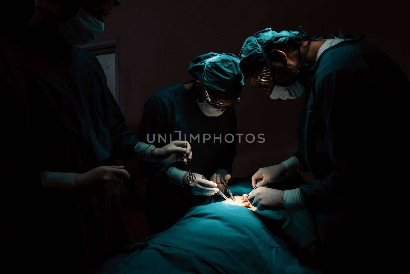 Surgical team performing surgery to patient in sterile operating room. In a surgery room lit by a lamp, a professional and confident surgical team provides medical care to an unconscious patient.