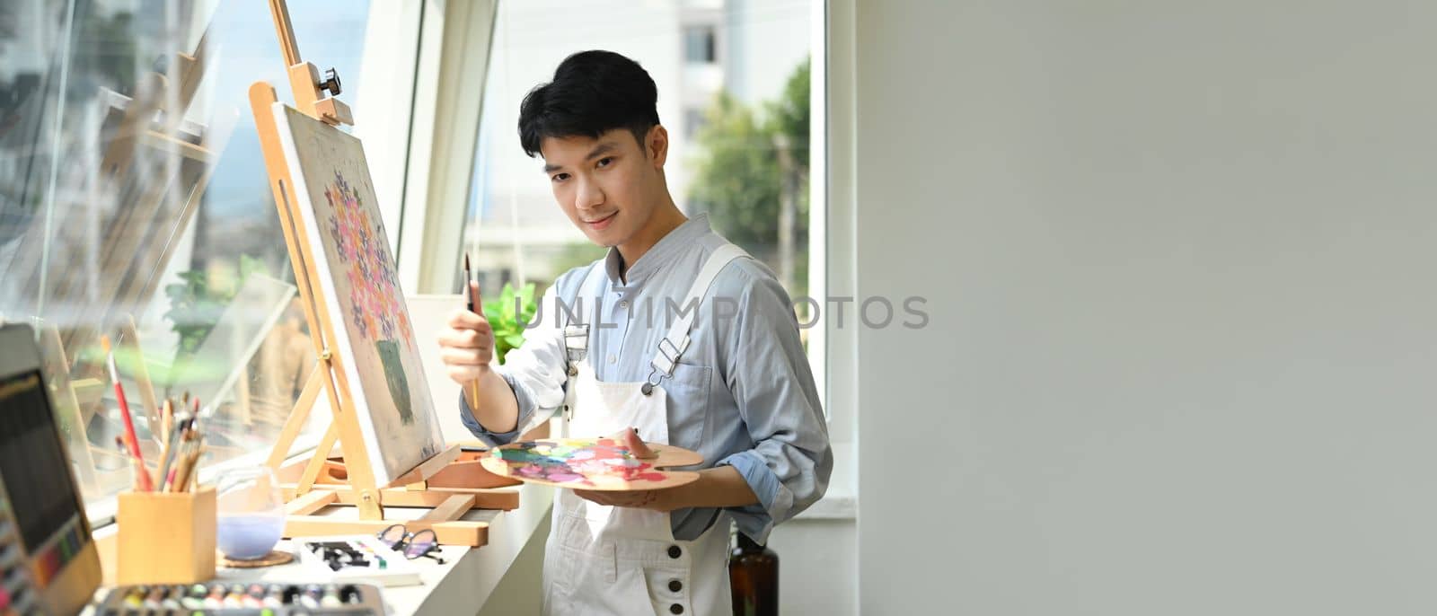 Asian man artist holding palette and paint brush standing in bright art studio. Panoramic web banner with copy space on the right by prathanchorruangsak