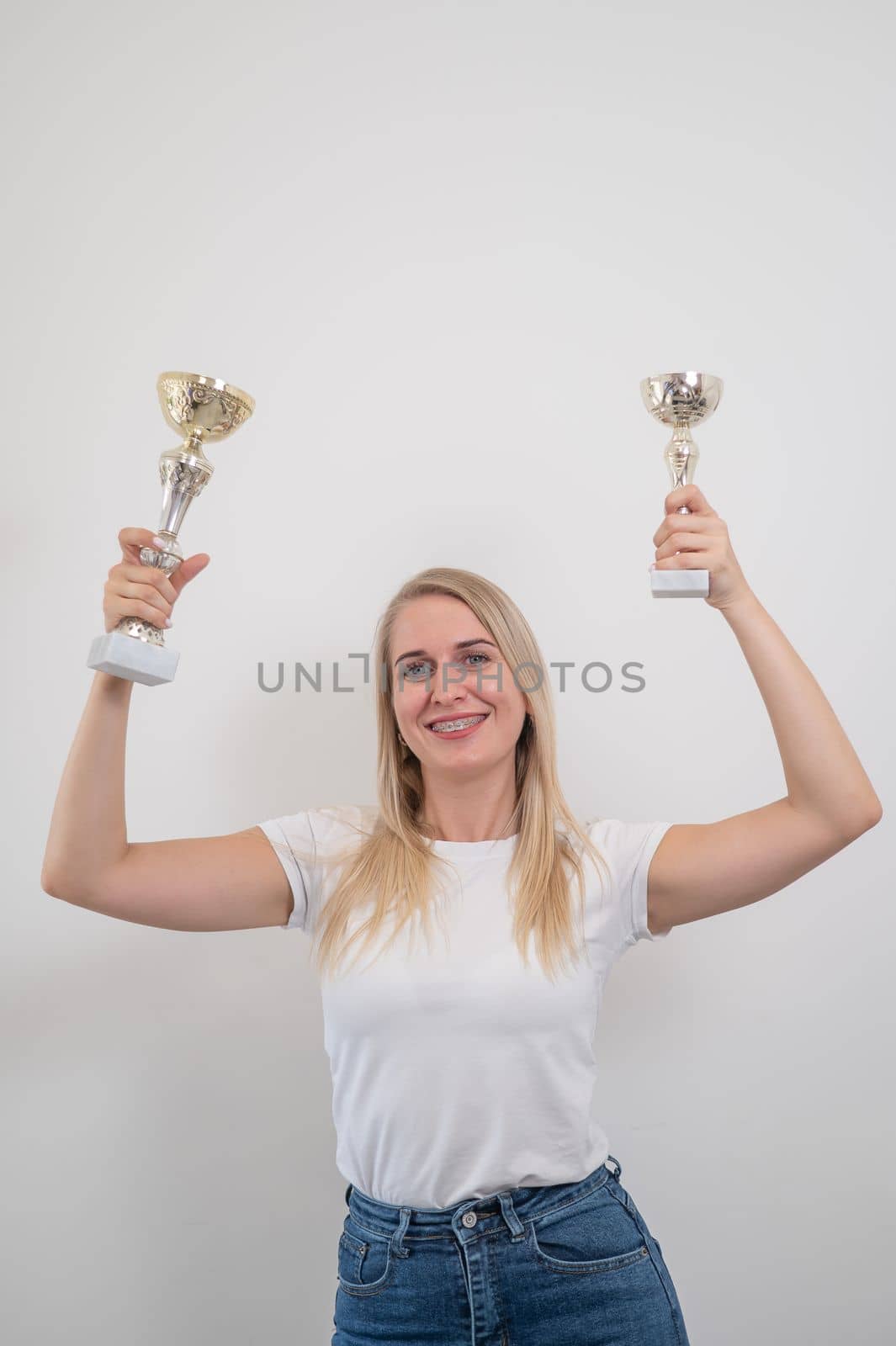 The blonde smiles and holds two golden trophy cup against the background of a white wall