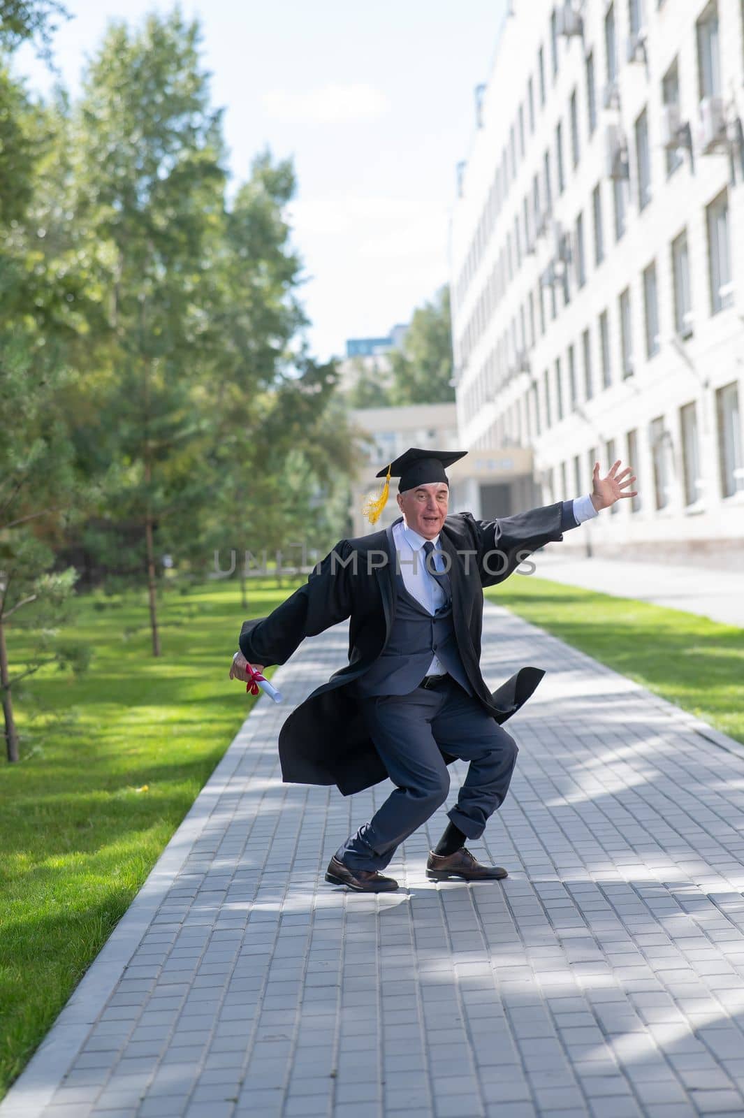 Old happy man in graduation gown jumping outdoors and holding diploma. Vertical