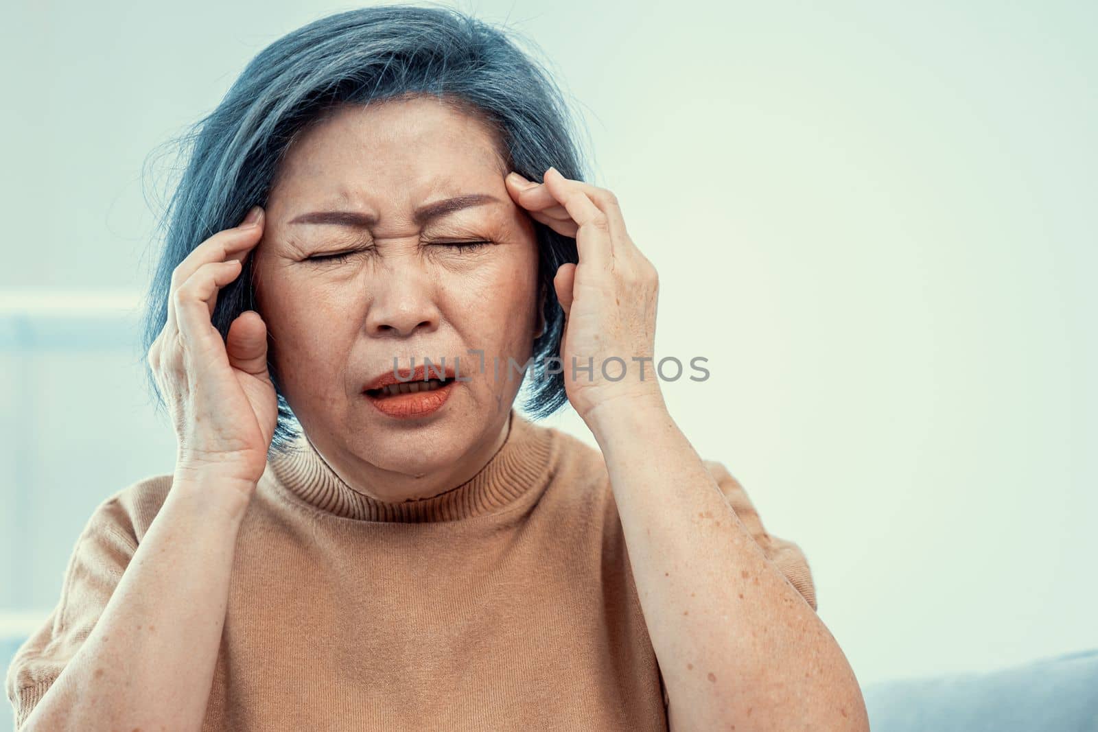 An agonizing senior woman with a headache, compress her temple with both hands at her contented living room. Senior health, nursing home, caretaker service.