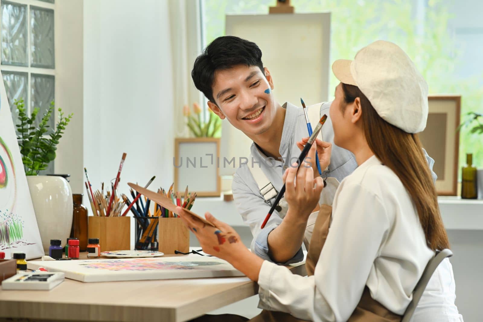 Asian man painting face his girlfriend during painting in at art studio. Leisure activity, creative hobby and art concept.