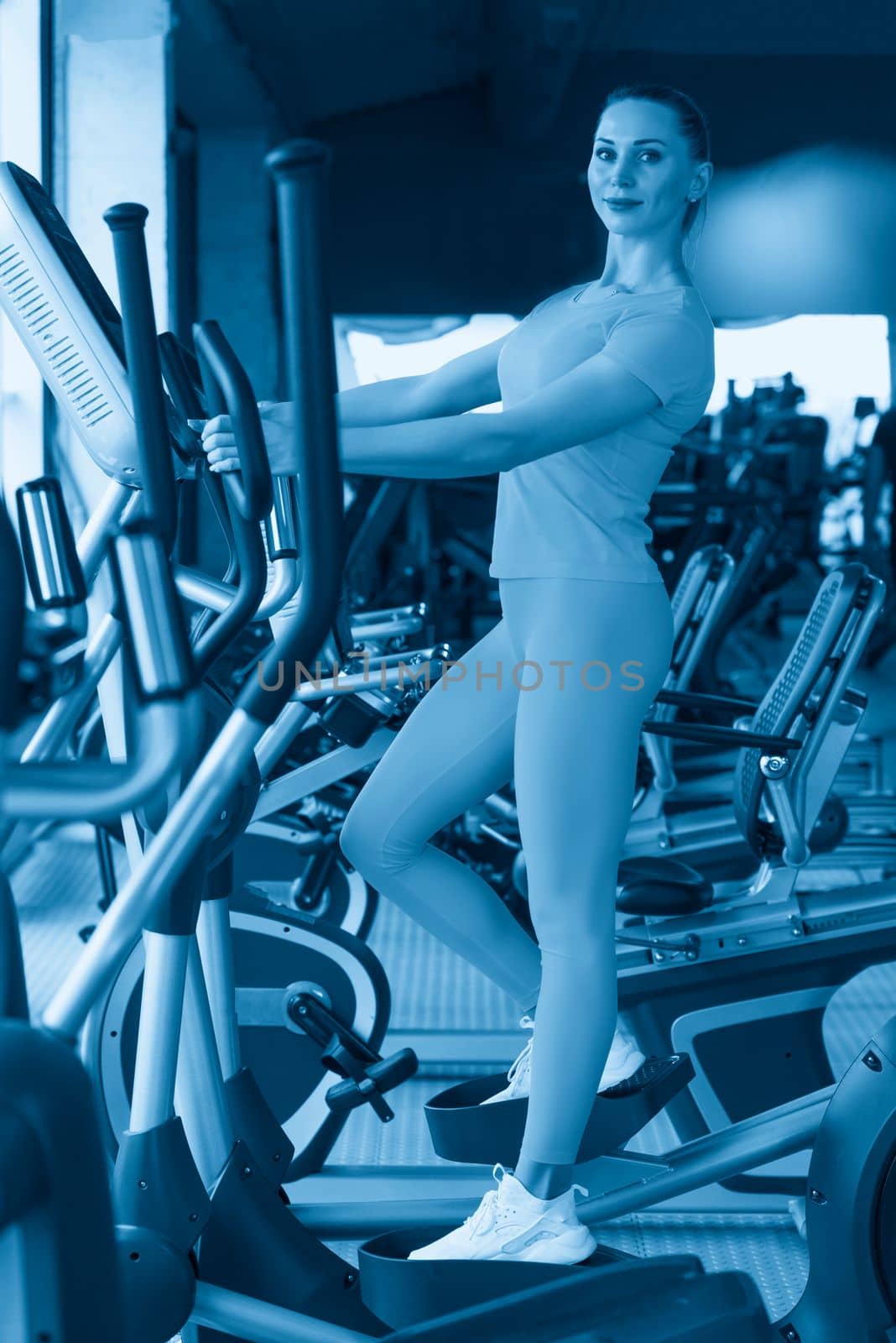 Young woman standing on Elliptical Machine in gym by Mariakray