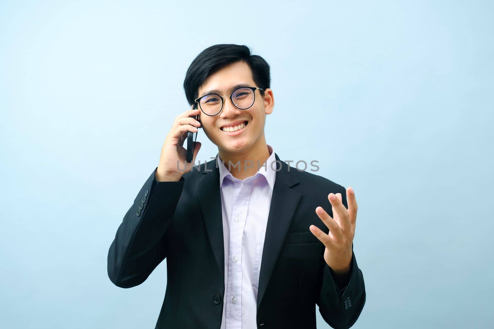 Portrait of young smart asian businessman in eyeglasses standing, smiling, and talking on mobile phone about business with light blue isolated background. Business and connection concept.
