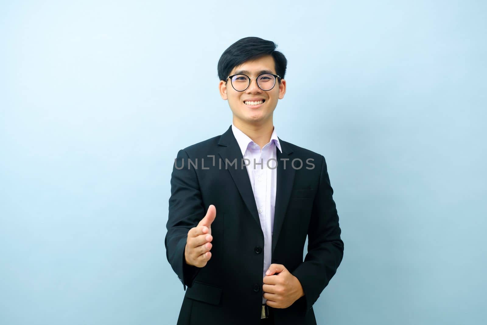 Portriat of asian young smart happy businessman dressed in suit standing straight, smiling, and pointing finger at camera with isolated light blue background. Business concept.