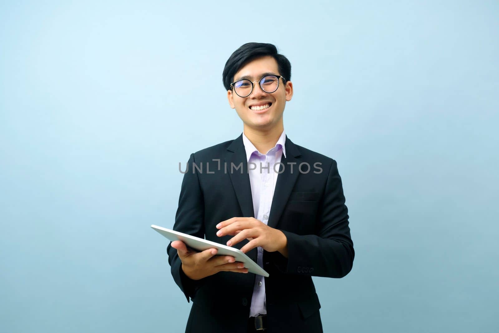 Portrait of young smart asian businessman wearing glasses standing and smiling while using tablet for working or communicating with light blue isolated background. Business, connection concept.
