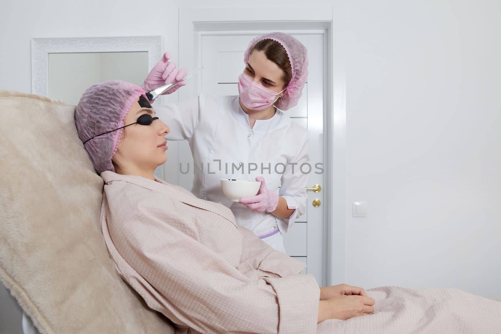 Carbon peeling in beauty salon. Cosmetologist applying black mask on the face of a beautiful woman for carbon peeling by Mariakray