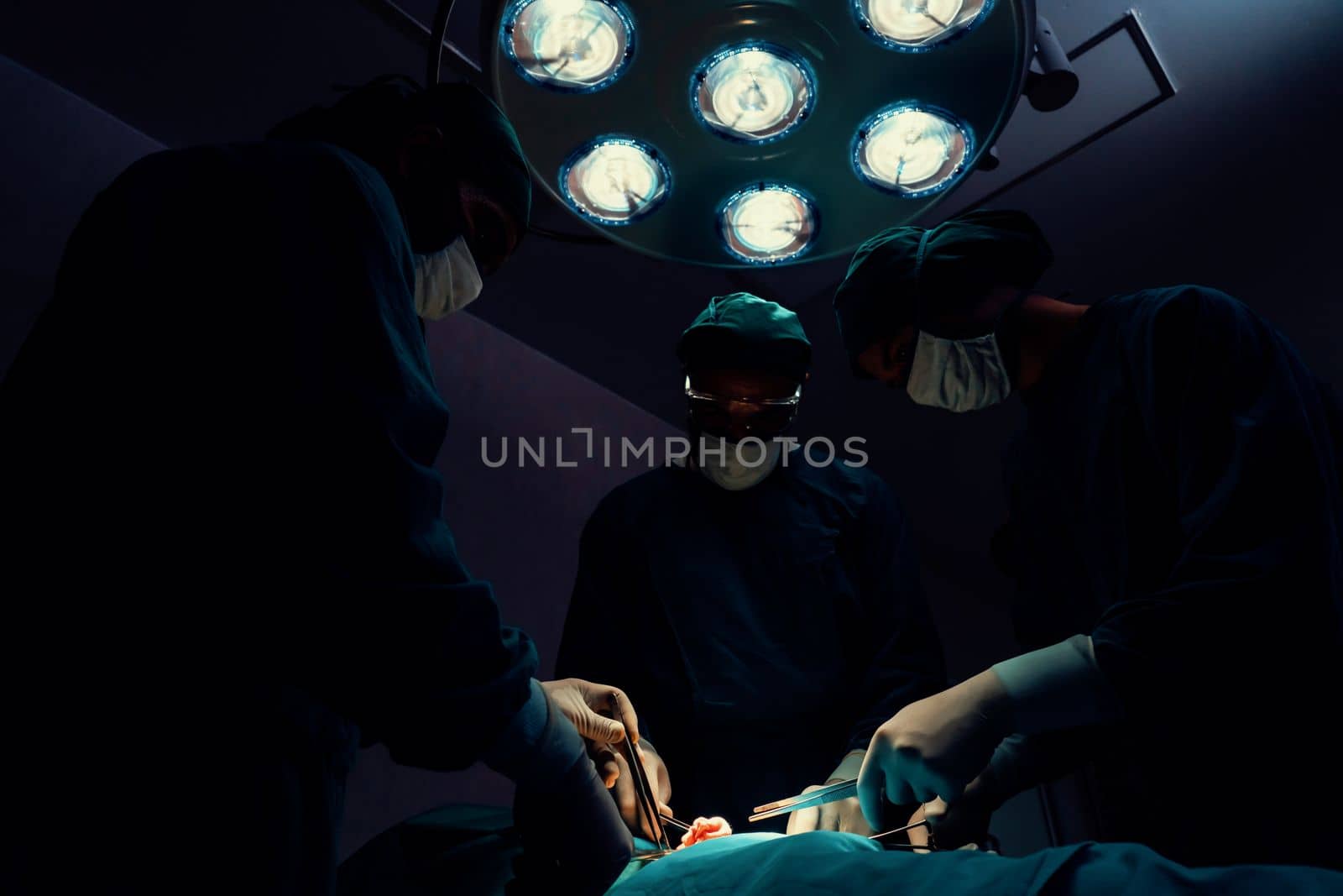 Surgical team performing surgery to patient in sterile operating room. In a surgery room lit by a lamp, a professional and confident surgical team provides medical care to an unconscious patient.