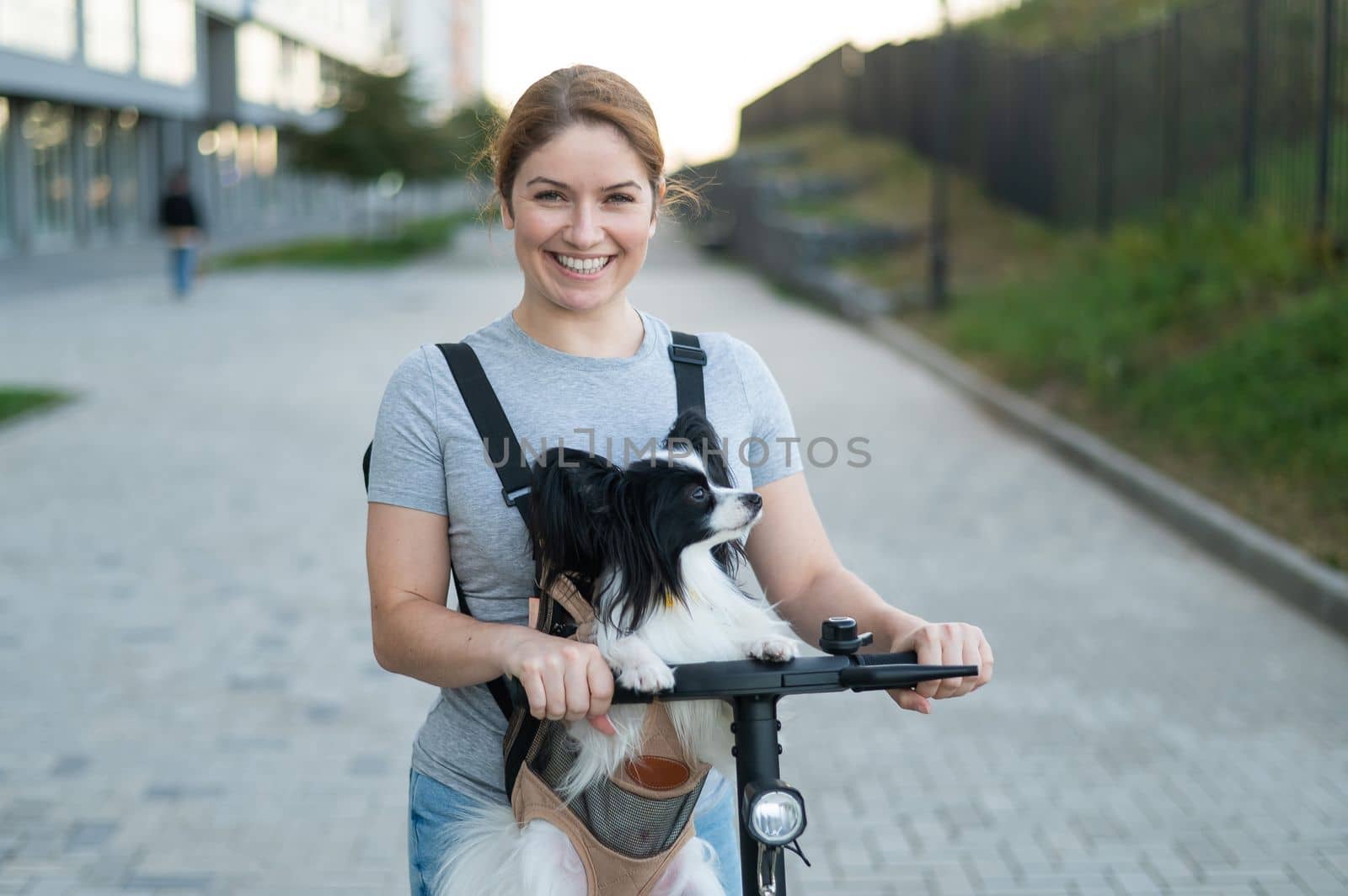 A woman rides an electric scooter with a dog in a backpack. Pappilion Spaniel Continental in a sling