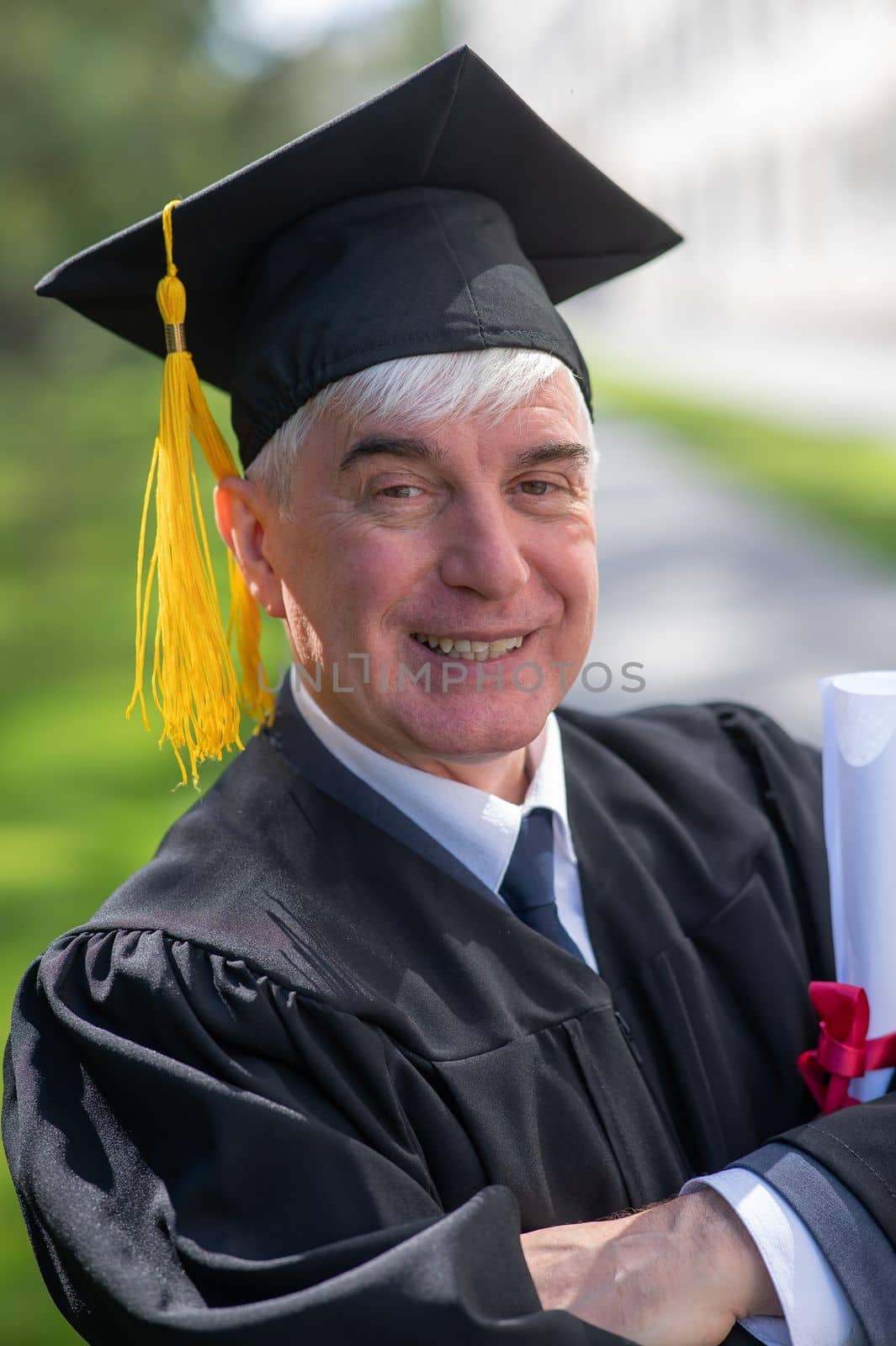 Portrait of an elderly man in a graduation gown and with a diploma in his hands outdoors. Vertical