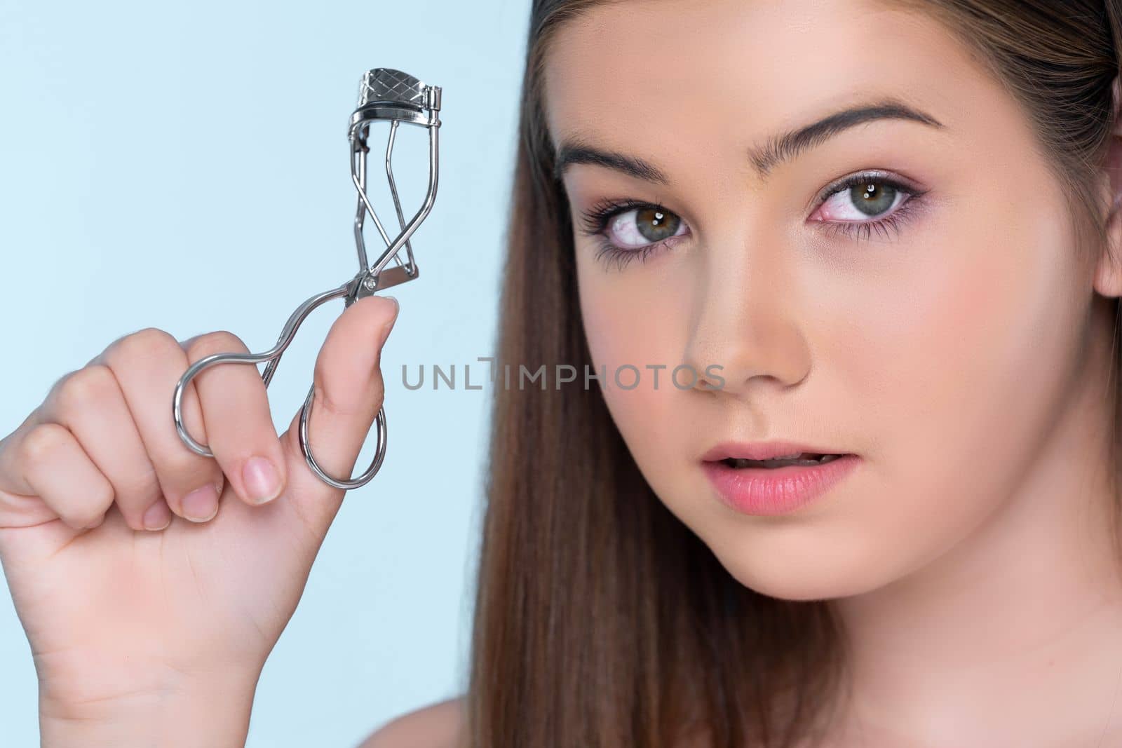 Closeup happy young charming brunette holding eyelash curler in her hand as makeup accessory. Beautiful girl applying eyelash curler in studio with isolated background for copyspace.