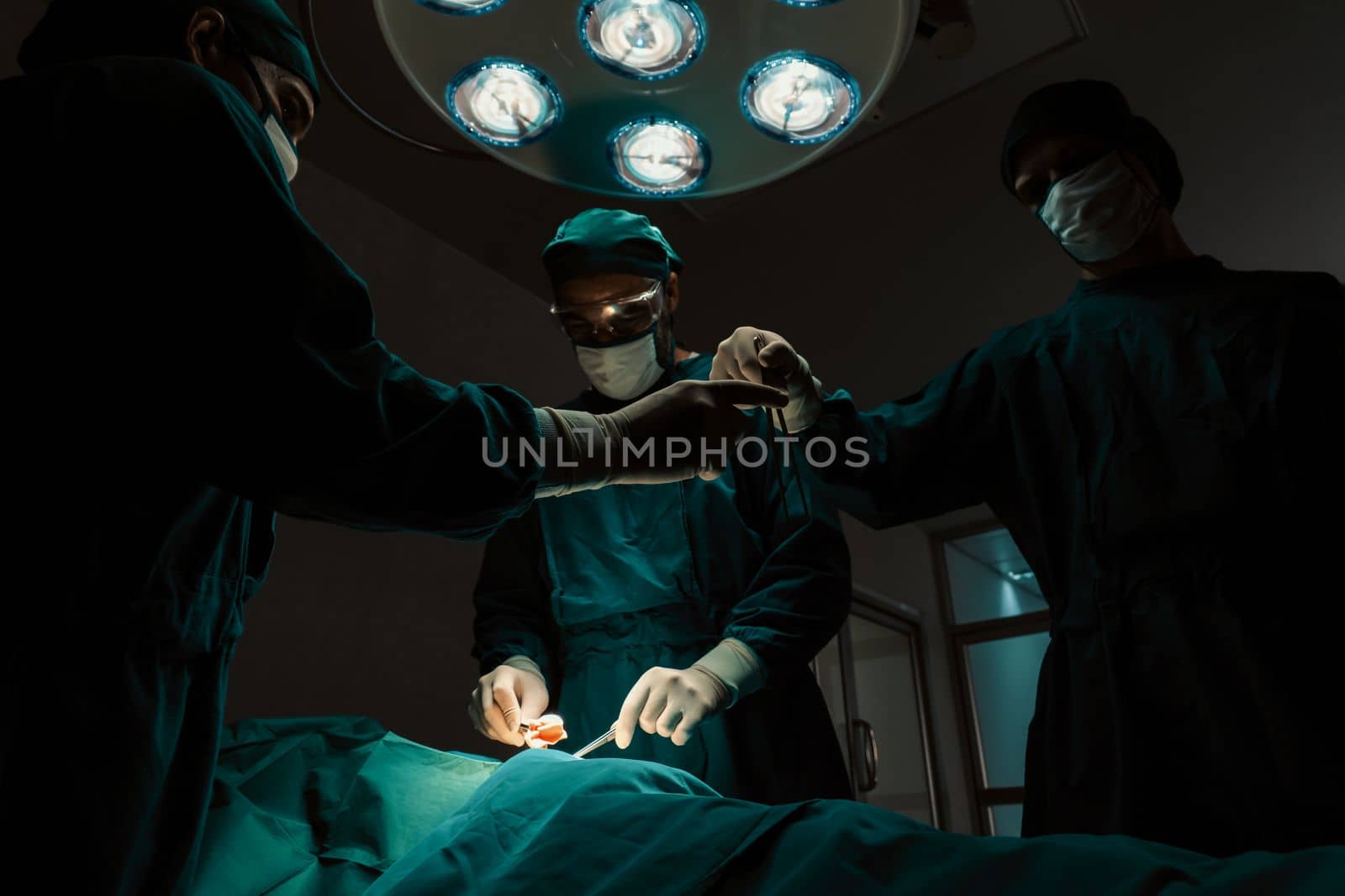 Surgical team performing surgery to patient in sterile operating room. In a surgery room lit by a lamp, a professional and confident surgical team provides medical care to an unconscious patient.