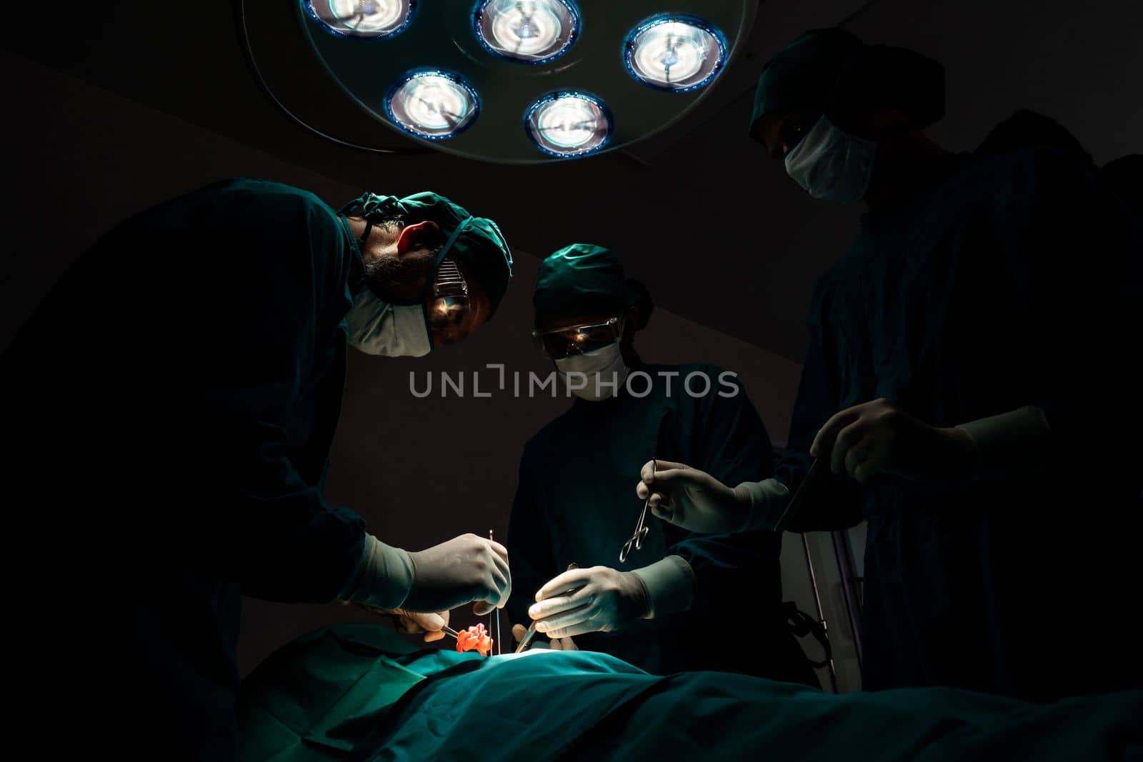 Surgical team performing surgery to patient in sterile operating room. In a surgery room lit by a lamp, a professional and confident surgical team provides medical care to an unconscious patient.