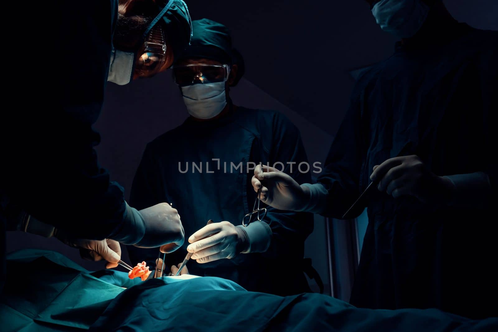 Surgical team performing surgery to patient in sterile operating room. In a surgery room lit by a lamp, a professional and confident surgical team provides medical care to an unconscious patient.