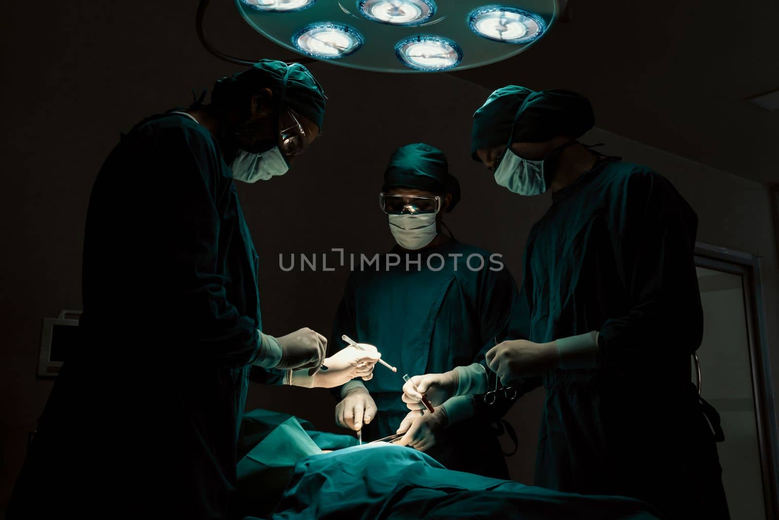 Surgical team performing surgery to patient in sterile operating room. In a surgery room lit by a lamp, a professional and confident surgical team provides medical care to an unconscious patient.