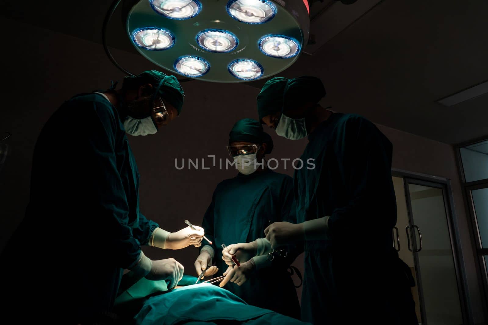 Surgical team performing surgery to patient in sterile operating room. In a surgery room lit by a lamp, a professional and confident surgical team provides medical care to an unconscious patient.