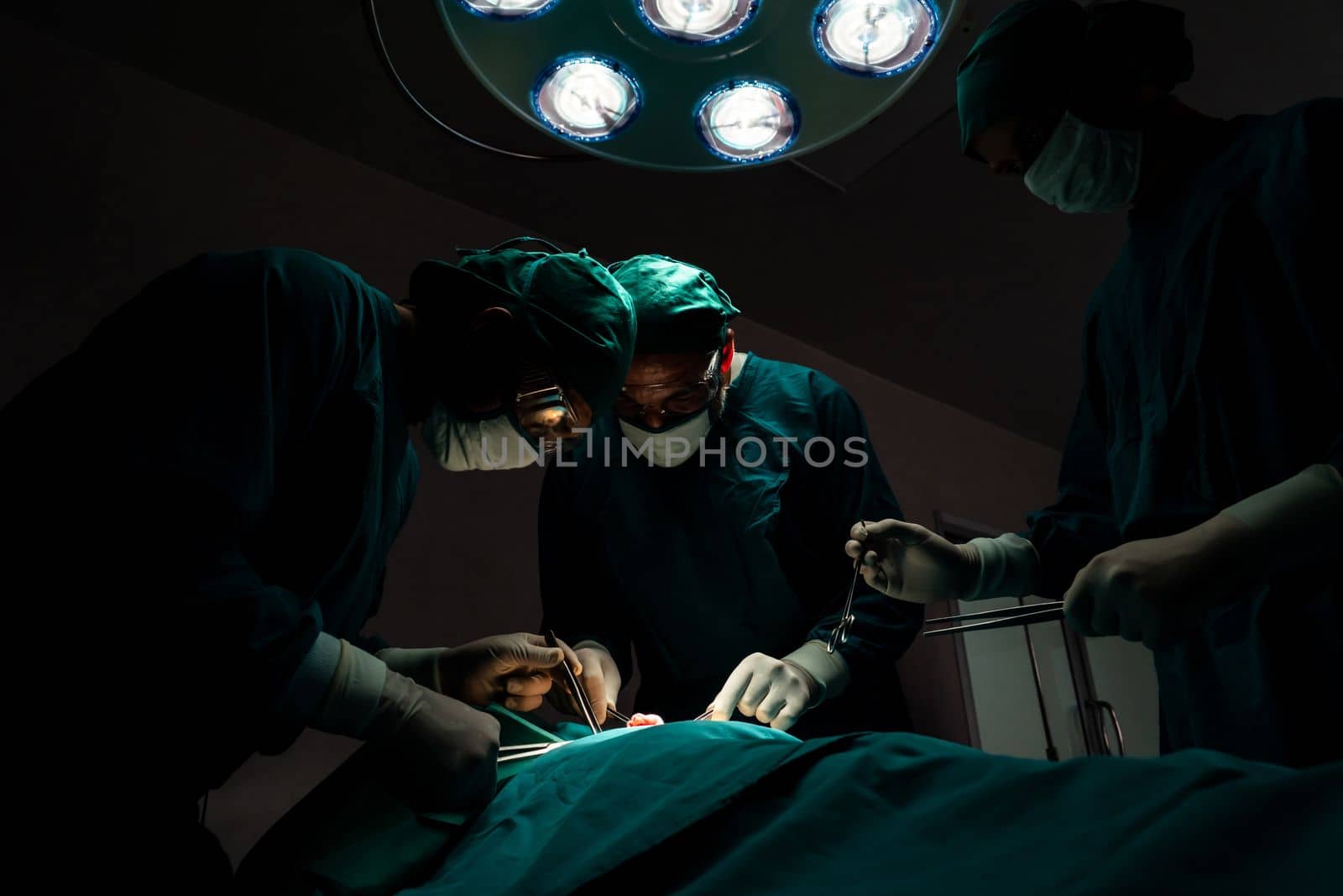 Surgical team performing surgery to patient in sterile operating room. In a surgery room lit by a lamp, a professional and confident surgical team provides medical care to an unconscious patient.