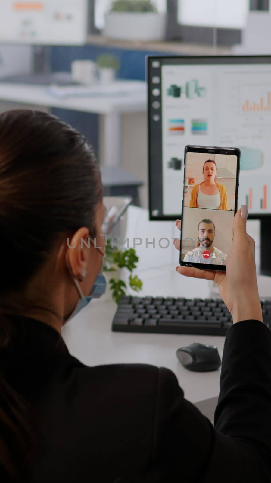 Executive business woman wearing protective face mask using phone for online videocall conference with remotely team. Coworkers working in background respecting social distance