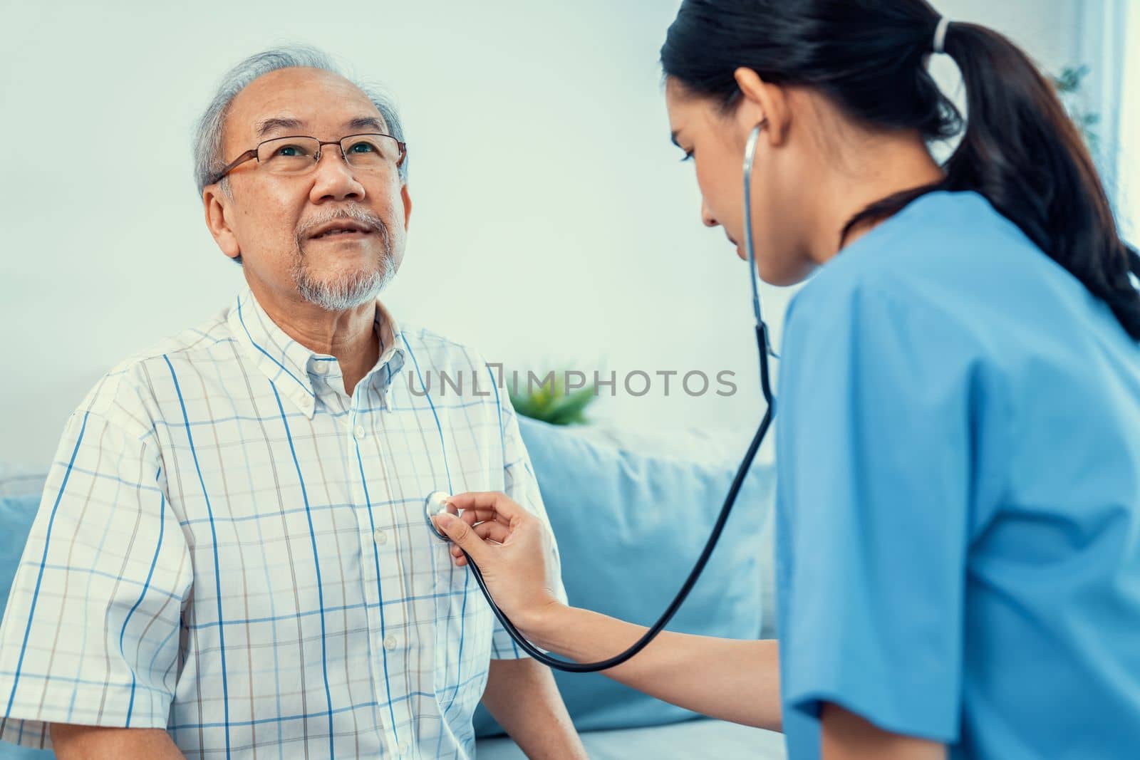 Caring young female doctor examining her contented senior patient with stethoscope in living room. Medical service for elderly, elderly sickness, declining health.