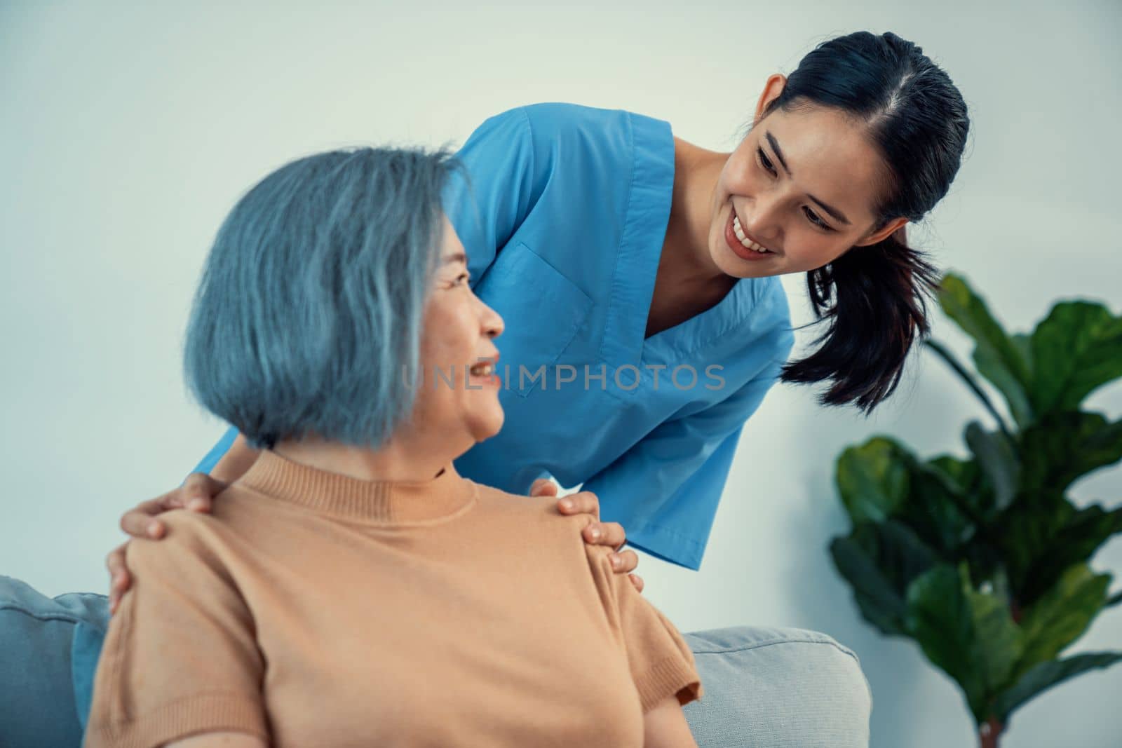 A caregiver rest her hands on the shoulders of a contented senior patient while she sitting on the sofa at home.