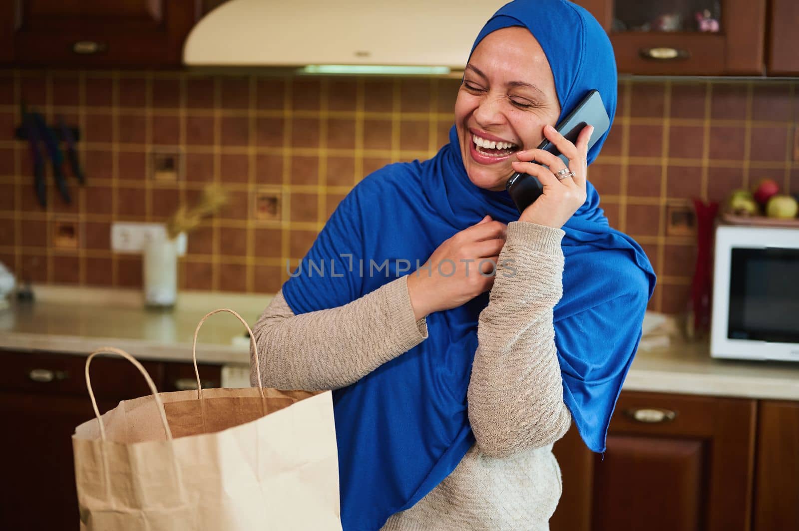 Stunning delightful Middle Eastern Muslim woman in a blue hijab, laughing, talking emotionally on mobile phone while unpacking shopping bag with groceries, standing in the cozy wooden kitchen at home