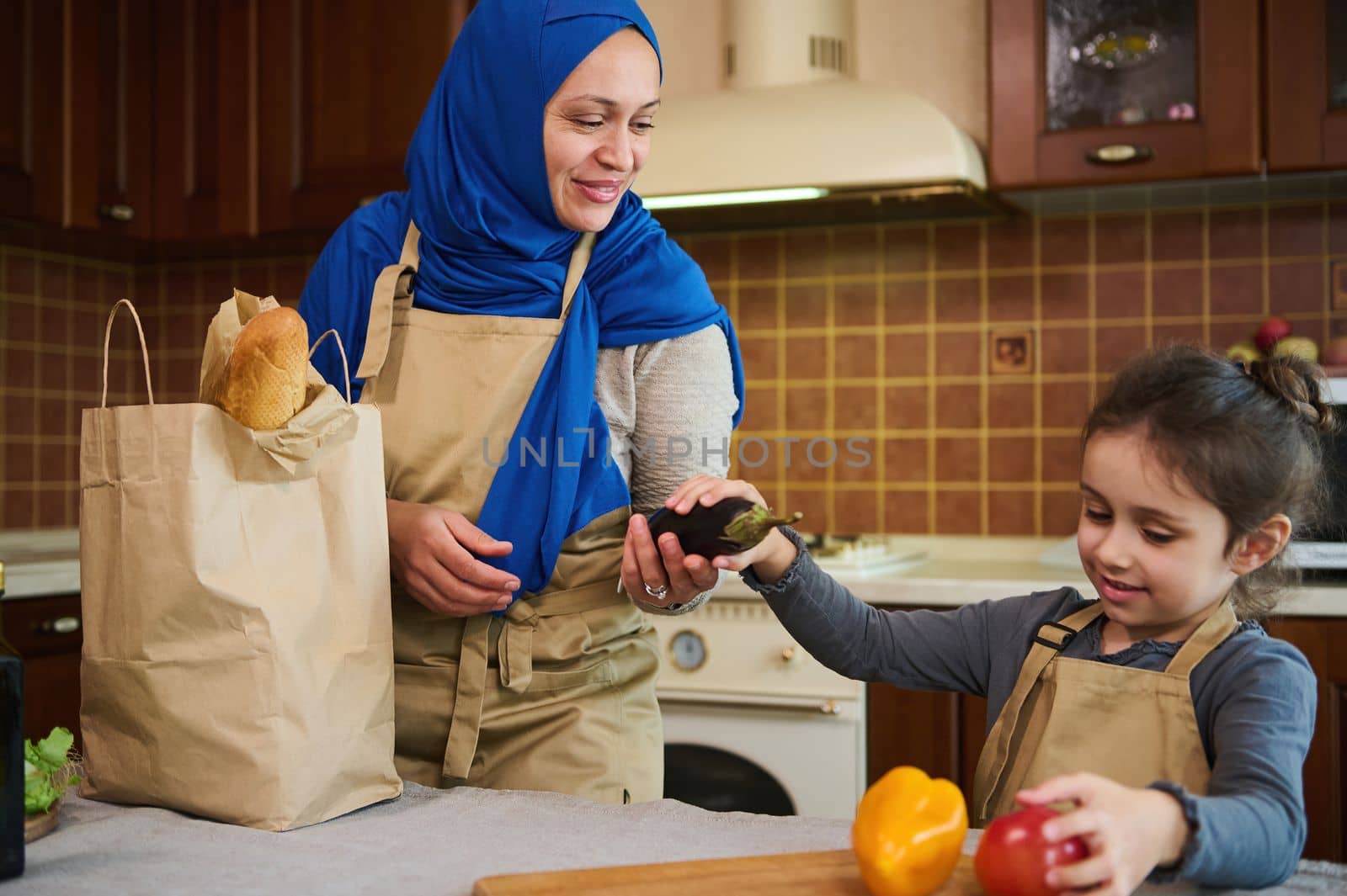 Cheerful happy Middle Eastern Muslim woman housewife in blue hijab, a loving mother and her lovely daughter unpacking grocery bag after shopping. Customers of online supermarkets. Food delivery