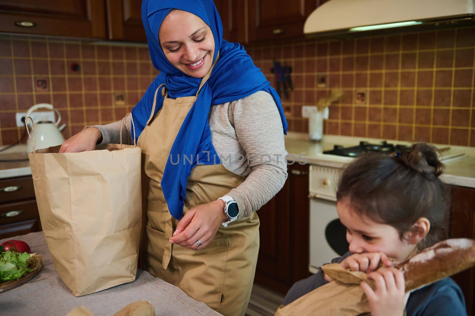 Beautiful Middle-Eastern Muslim woman with head covered in hijab, unpacking groceries with her lovely daughter, when arriving from grocery shopping. Food delivery. Ethical consumerism. Eco packaging