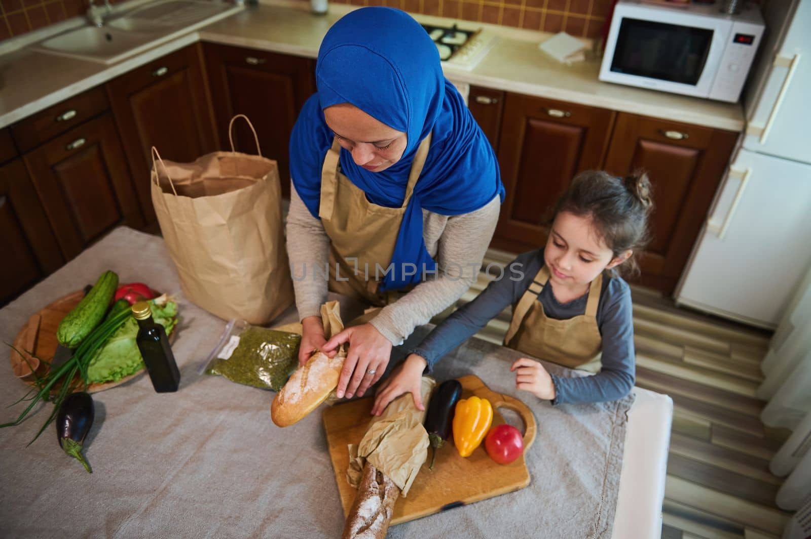 Top view. Happy Middle Eastern family relationship at home. Arab Muslim woman, loving mom with little daughter unpacking food, vegetable and fruit from supermarket bag together for preparing meal