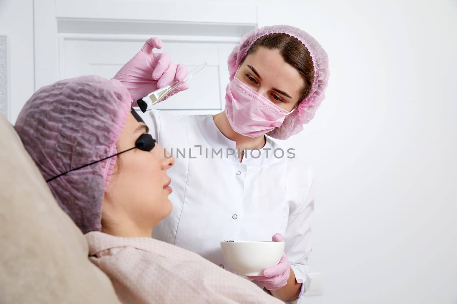 Carbon peeling in beauty salon. Cosmetologist applying black mask on the face of a beautiful woman for carbon peeling by Mariakray
