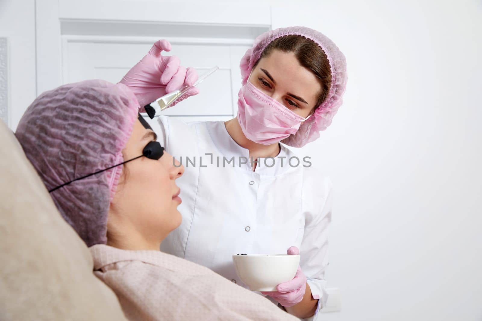 Carbon peeling in beauty salon. Cosmetologist applying black mask on the face of beautiful woman for carbon peeling