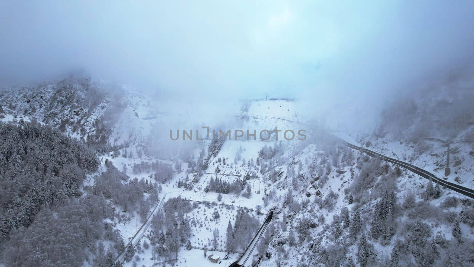 Snowy mountains with coniferous trees in the clouds. Medeo Dam. Everything is in fog and snow. Christmas and New Year have come. Aerial view from the drone on road, dam and trees. Almaty, Kazakhstan