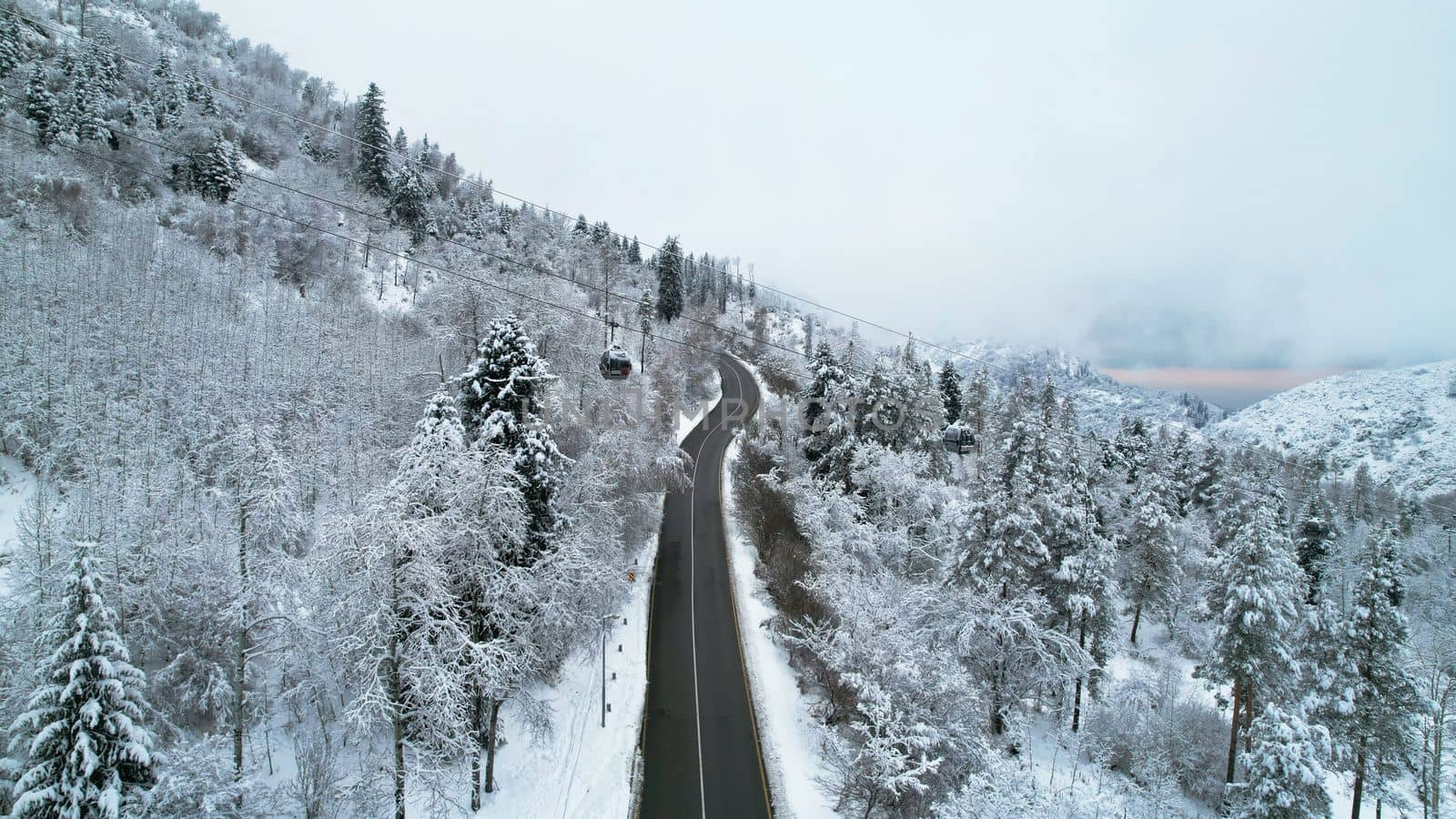 Cable car in the mountains in the winter forest. The cabins move along the gondola road through the winter snow forest and fog. White clouds covered the mountains. Top view from a drone. Medeo, Almaty