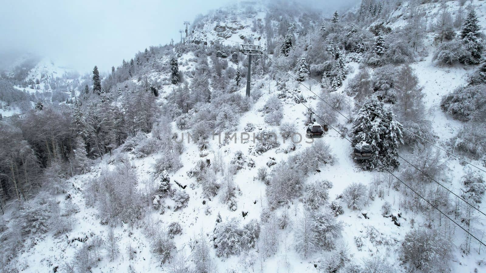 Cable car in the mountains in the winter forest. The cabins move along the gondola road through the winter snow forest and fog. White clouds covered the mountains. Top view from a drone. Medeo, Almaty