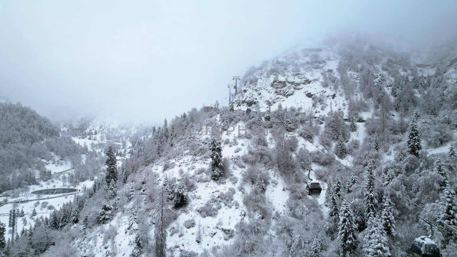 Cable car in the mountains in the winter forest. The cabins move along the gondola road through the winter snow forest and fog. White clouds covered the mountains. Top view from a drone. Medeo, Almaty