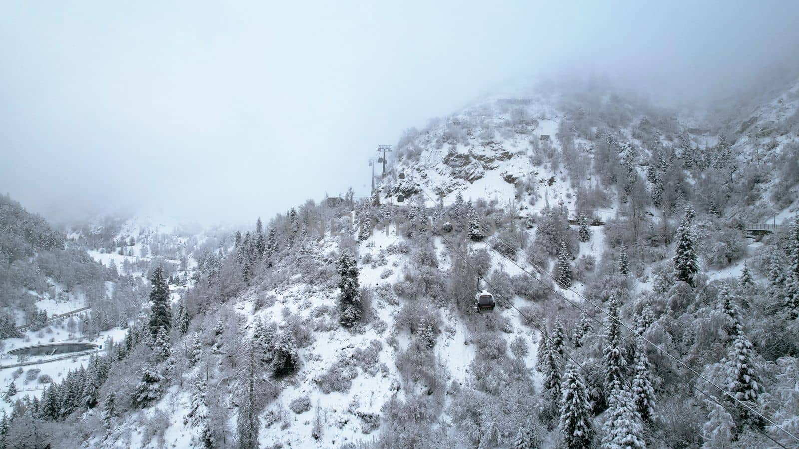 Cable car in the mountains in the winter forest. The cabins move along the gondola road through the winter snow forest and fog. White clouds covered the mountains. Top view from a drone. Medeo, Almaty