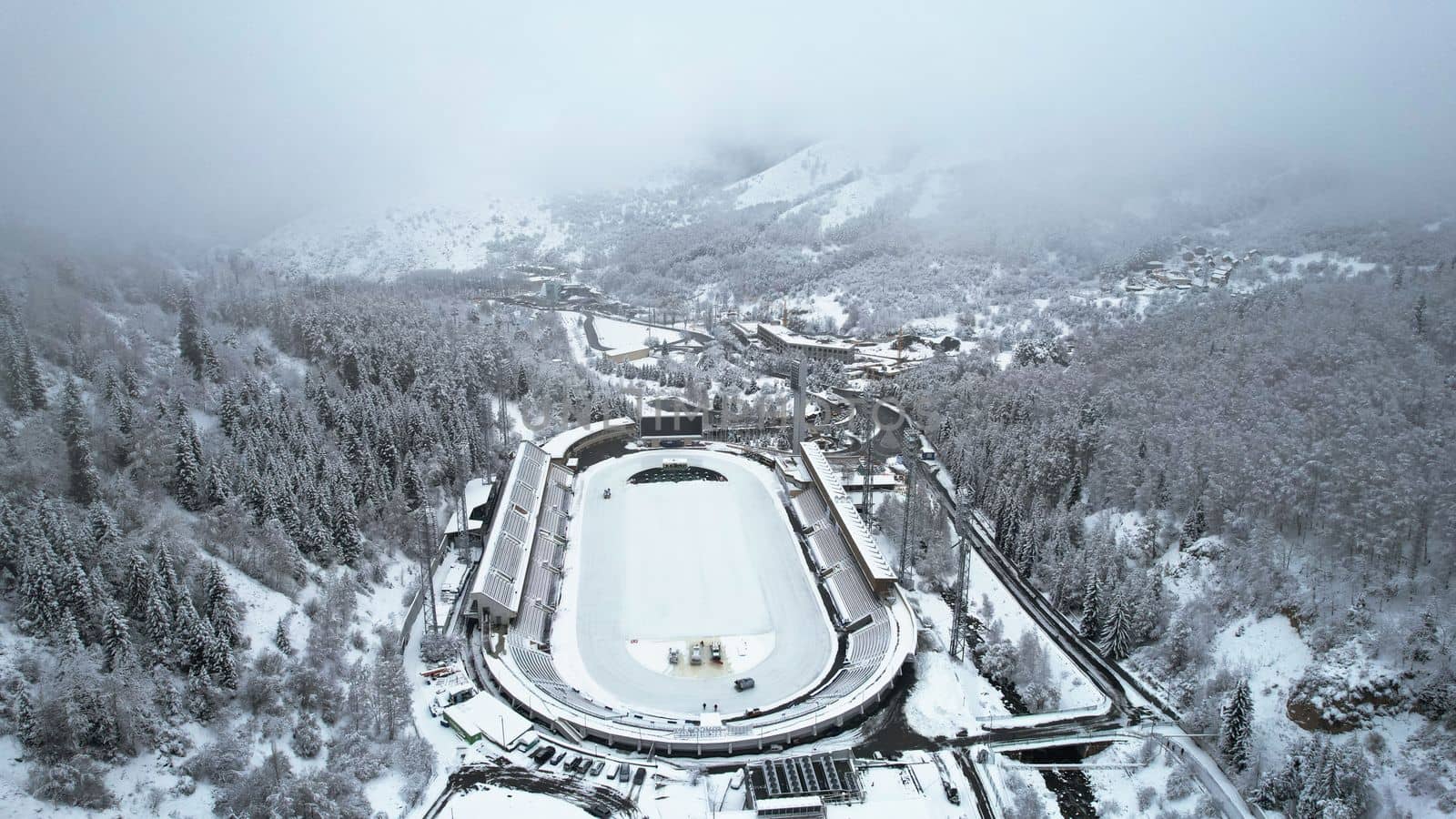 Winter alpine skating rink Medeo in the mountains. Drone view of the snowy forest and mountains. White clouds and fog in the gorge. Snow removal is underway. A large stadium. Almaty, Kazakhstan