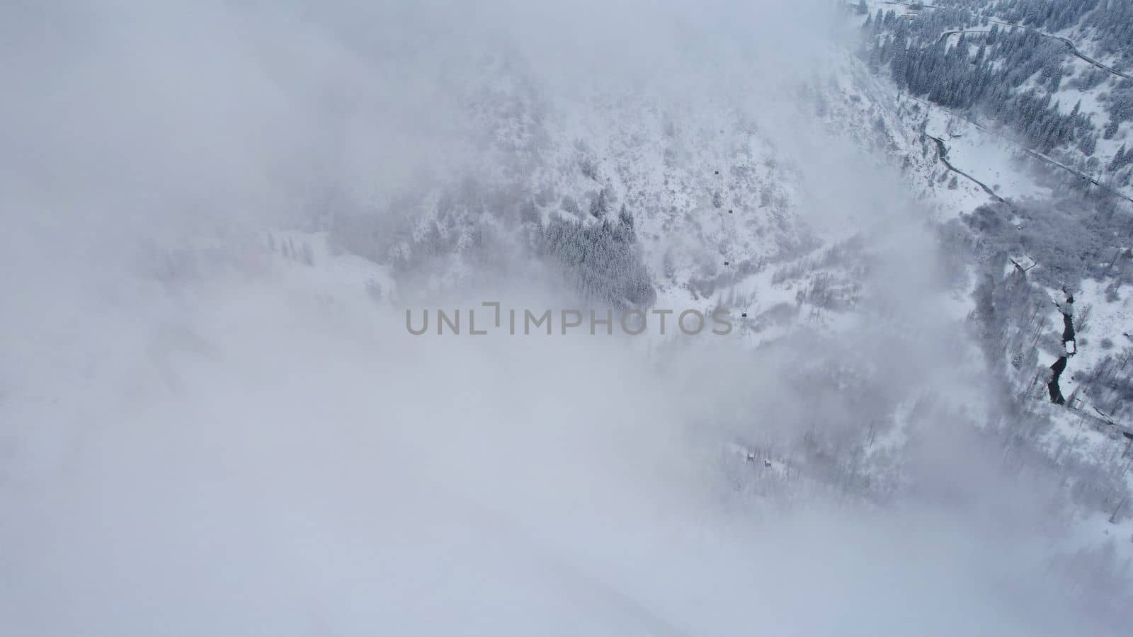 Snowy mountains with coniferous trees in the clouds. Medeo Dam. Everything is in fog and snow. Christmas and New Year have come. Aerial view from the drone on road, dam and trees. Almaty, Kazakhstan