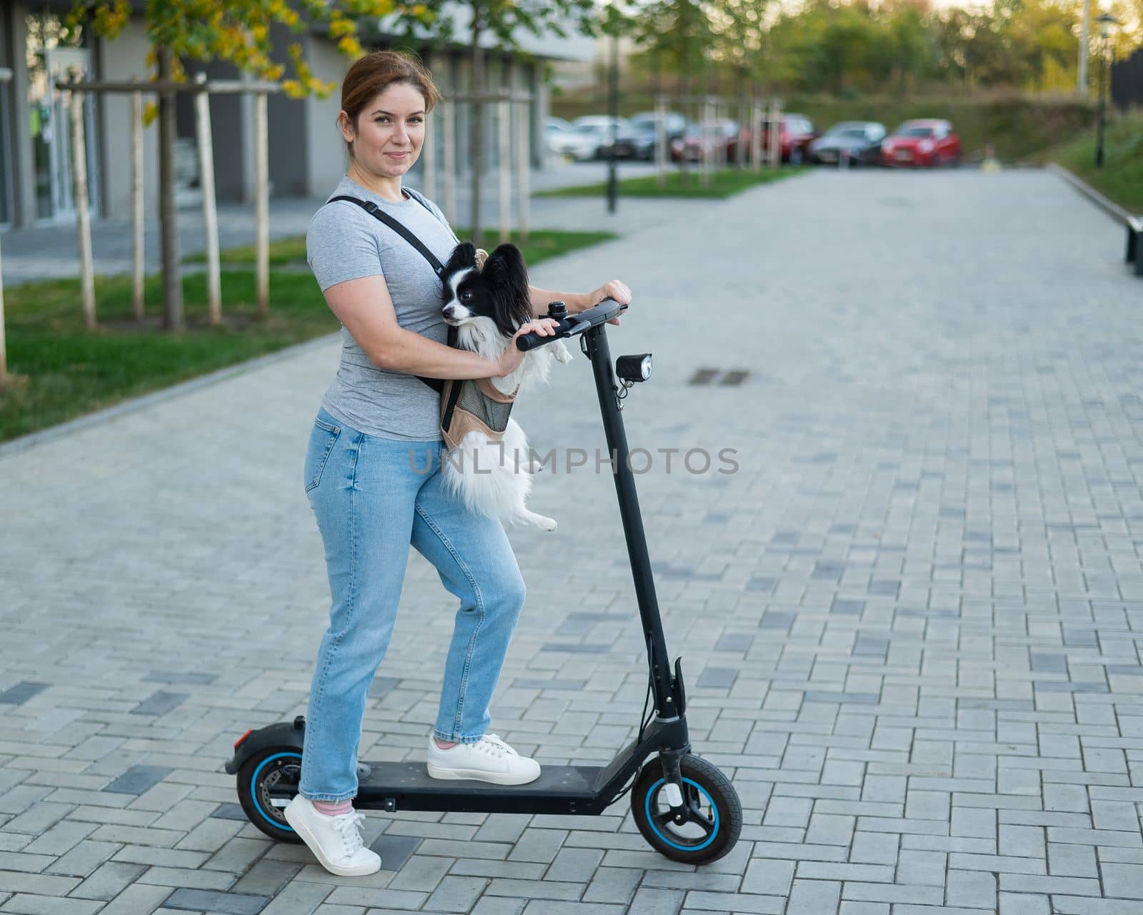 A woman rides an electric scooter with a dog in a backpack. Pappilion Spaniel Continental in a sling