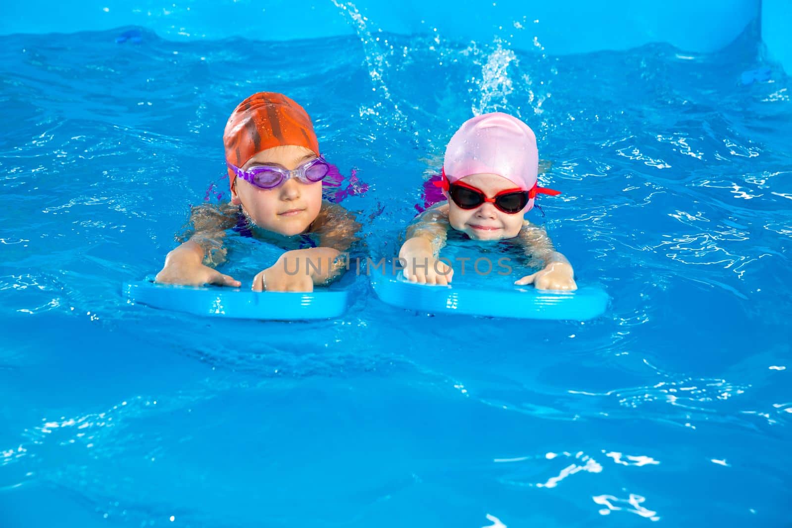 Two little girls learning how to swim in swimming pool using flutter boards by Mariakray