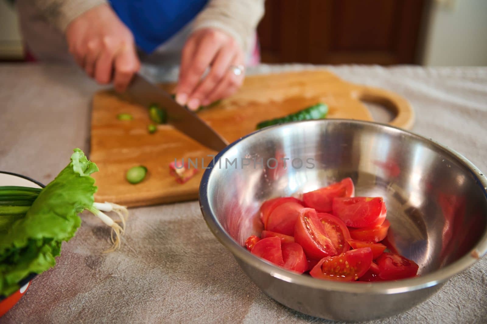 Selective focus on a metal bowl with chopped ripe juicy red tomatoes on a kitchen table with linen tablecloth, against a blurred background of woman's hands slicing cucumbers on a wooden cutting board